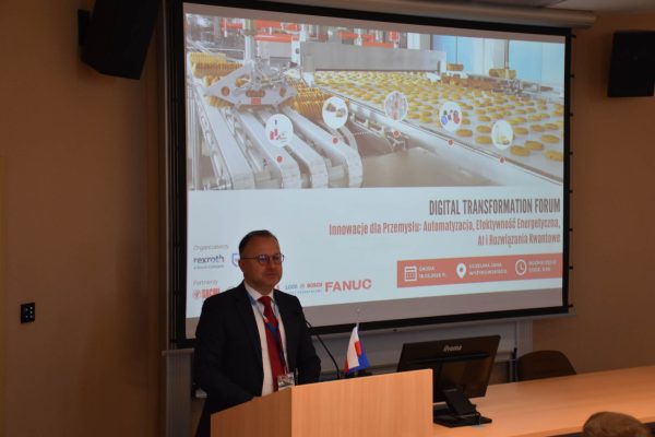 Prelegent przy mównicy na tle planszy Digital Transformation Forum z robotami przemysłowymi.