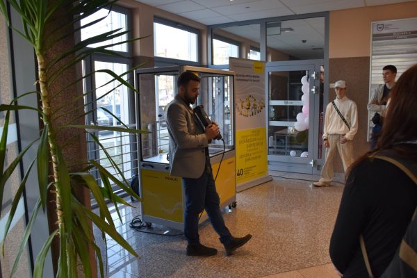 Prelegent z mikrofonem prowadzi pokaz przy stanowisku z robotem przemysłowym.