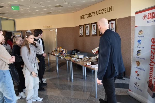 Prowadzący prezentuje wyposażenie stołu pokazowego uczniom w holu uczelni.