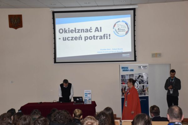 Prowadzący na scenie pod slajdem „Okiełznać AI - uczeń potrafi!”.