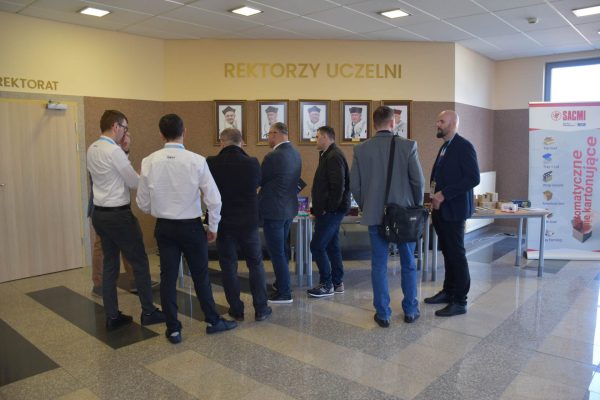 Uczestnicy konferencji rozmawiają przy stanowisku w holu pod portretami rektorów.