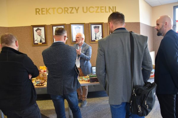 Grupa uczestników słucha mężczyzny prezentującego materiały przy stole pokazowym.
