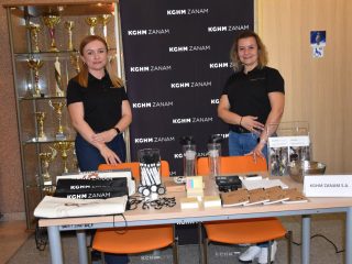 Dwie kobiety przy stoisku KGHM ZANAM z materiałami promocyjnymi i modelami urządzeń.
