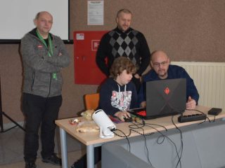 Cztery osoby przy stanowisku z laptopem i sprzętem elektronicznym w sali wykładowej.