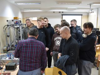 Grupa uczestników skupiona wokół stanowiska z urządzeniami w pracowni technicznej.