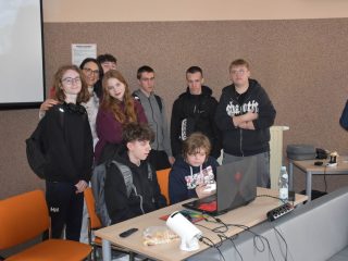 Grupa uczestników przy laptopie na stanowisku pokazowym UJW Whoop Racing Team.