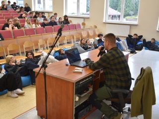 Prowadzący z mikrofonem przy biurku, otoczony uczestnikami siedzącymi na krzesłach i pufach.