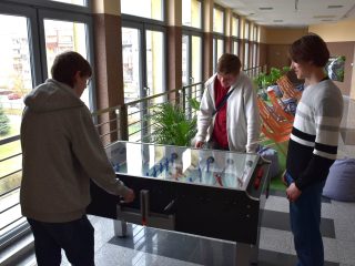 Trzej uczestnicy grają w piłkarzyki na korytarzu przy oknach.