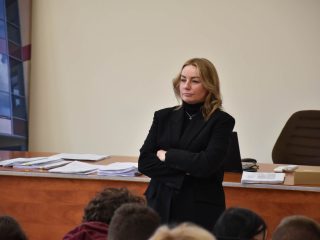 Prowadząca w czarnej marynarce przemawia przy biurku w sali wykładowej.