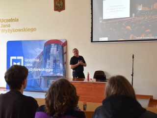 Prowadzący w czarnej koszuli prezentuje slajd uczestnikom spotkania w sali wykładowej.