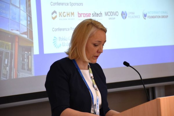 Prowadząca odczytuje tekst przy mikrofonie na tle planszy sponsorów konferencji.