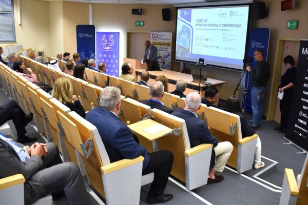 Widok sali konferencyjnej z publicznością, ekranem i kamerą przy przejściu.
