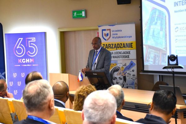Prelegent w szarym garniturze zwraca się do publiczności z przodu sali.
