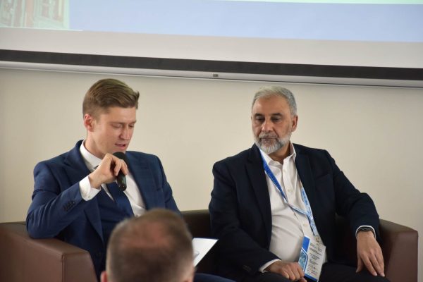 Moderator mówi do mikrofonu z notatkami w dłoni, obok siedzi brodaty panelista.