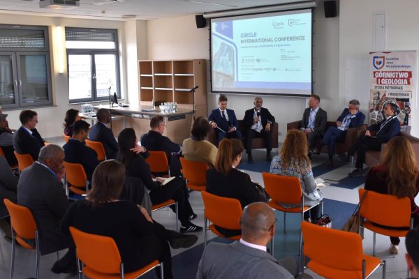 Widok sali z publicznością i pięcioma panelistami pod ekranem konferencji.