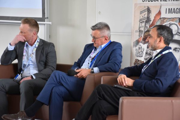 Trzej paneliści siedzą w fotelach, jeden trzyma mikrofon na kolanach.