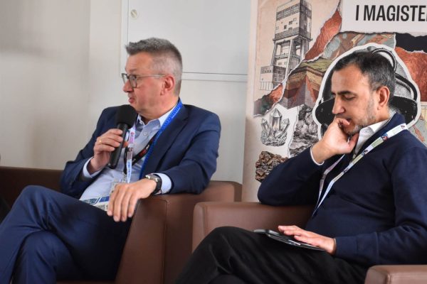 Panelista w niebieskim garniturze przemawia do mikrofonu, obok słucha drugi uczestnik.