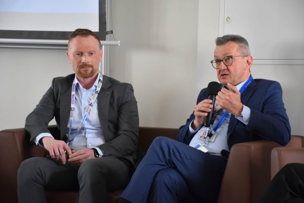 Panelista w niebieskim garniturze gestykuluje podczas wypowiedzi, obok siedzi drugi mówca.