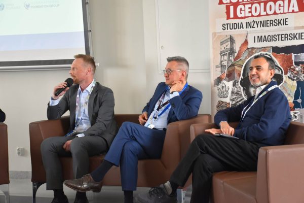 Panelista w szarej marynarce przemawia, dwaj pozostali uczestnicy słuchają.