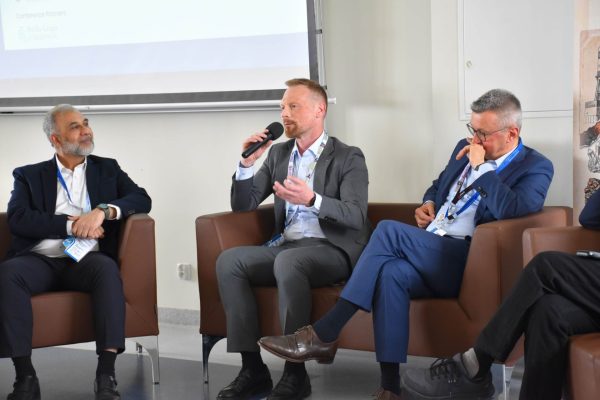 Mówca w szarej marynarce z mikrofonem podczas dyskusji w fotelach.
