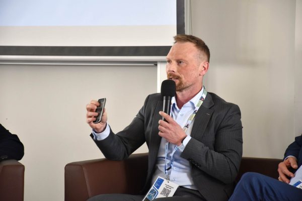Panelista w szarej marynarce prezentuje niewielkie urządzenie, trzymając mikrofon.