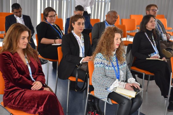 Uczestniczki konferencji z notesami siedzą na pomarańczowych krzesłach.