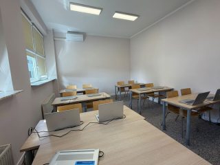 Sala zajęciowa z ławkami i laptopami przy stanowisku prowadzącego.