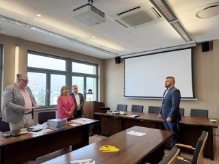 Absolwent w granatowym garniturze stoi przed trzyosobową komisją w sali UJW.