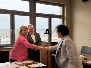 Kobieta w różowej marynarce gratuluje absolwentce w jasnoszarej marynarce uściskiem dłoni.