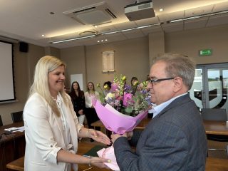 Kobieta w jasnym garniturze przekazuje bukiet mężczyźnie w okularach podczas podziękowań po obronach.