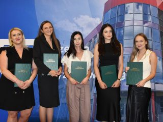 Pięć kobiet po obronach prac dyplomowych, na tle ścianki UJW; cztery trzymają zielone teczki.