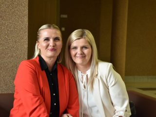 Dwie kobiety w czerwonej i kremowej marynarce pozują na korytarzu uczelni.