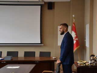 Mężczyzna w granatowym garniturze stoi przy stole komisji podczas obron zarządzania.