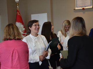 Uczestniczki obron zarządzania rozmawiają w sali; kobieta w białej bluzce trzyma zieloną pracę.