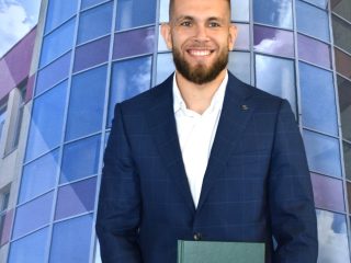 Uśmiechnięty absolwent w granatowym garniturze prezentuje zieloną pracę licencjacką.