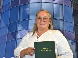 Zbliżenie absolwentki w okularach i białej bluzce prezentującej zieloną pracę licencjacką.