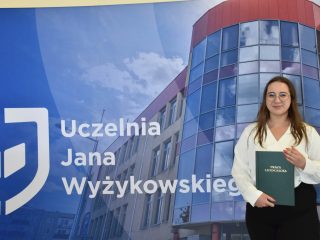 Absolwentka w okularach, białej bluzce i czarnych spodniach trzyma pracę licencjacką na tle fotografii uczelni.