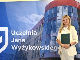 Absolwentka w kremowym garniturze prezentuje zieloną pracę licencjacką.