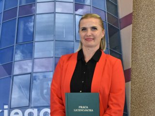 Absolwentka w czerwonej marynarce trzyma zieloną pracę magisterską na tle fotografii UJW.