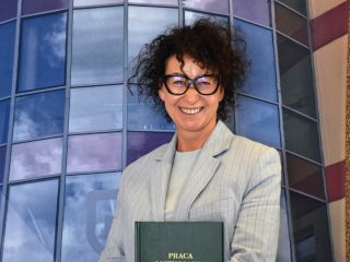 Uśmiechnięta absolwentka w okularach i jasnoszarej marynarce prezentuje zieloną pracę dyplomową.