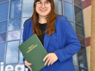 Absolwentka w niebieskim garniturze trzyma zieloną pracę dyplomową na tle fotografii budynku UJW.
