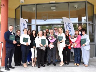 Grupa absolwentów i pracowników UJW przed wejściem do uczelni, z oprawionymi pracami i bukietami kwiatów.