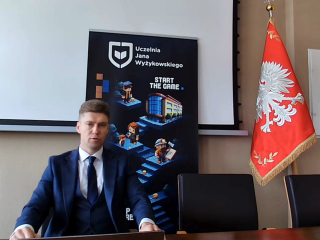 Mężczyzna w garniturze siedzi przy stole na tle planszy UJW i flagi Polski.