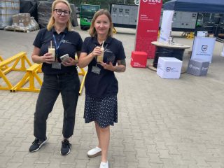 Dwie uczestniczki z napojami stoją na placu przed stoiskami.